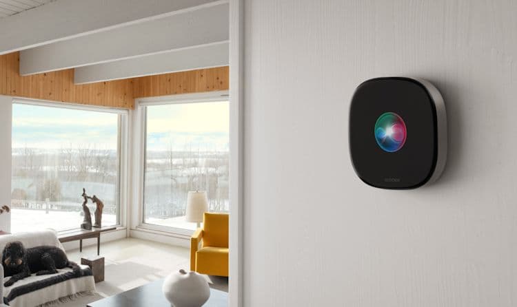 The Ecobee Smart Thermostat Premium mounted on a light-colored wall, with a smartphone on the left showing the ecobee app interface in a bright modern room.